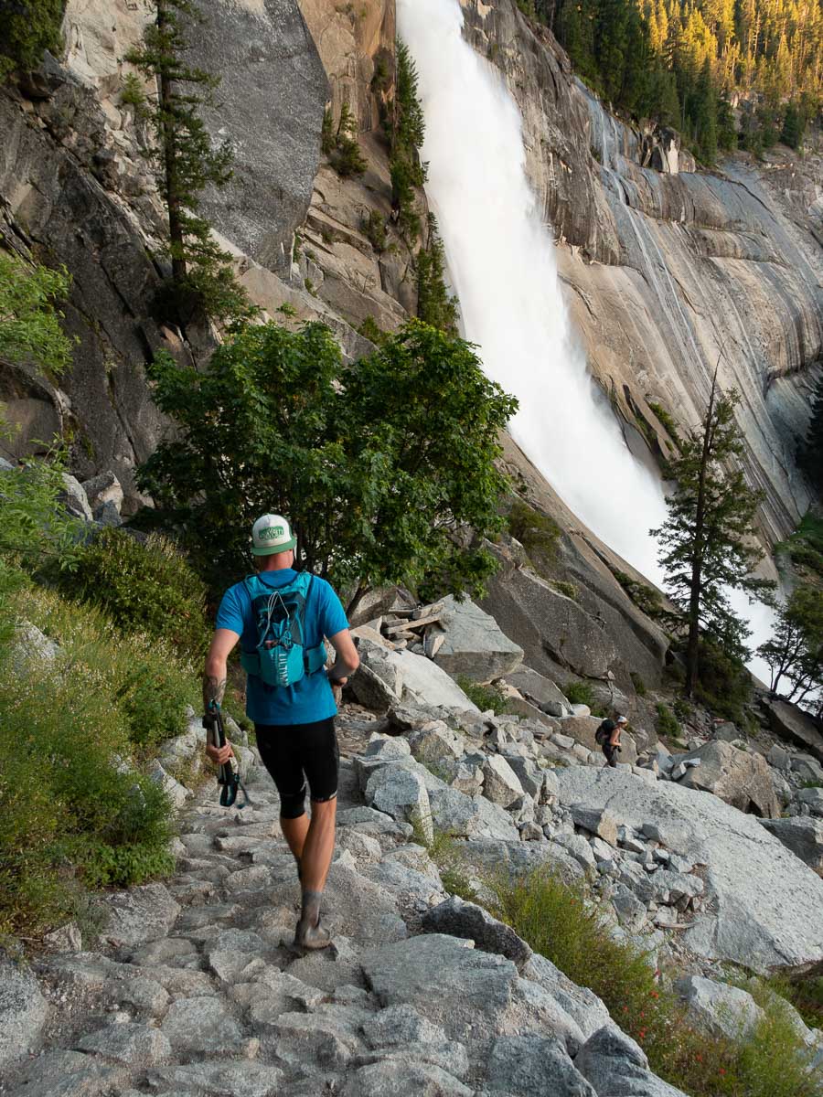 a man running down a rocky trail with a waterfall in the background.